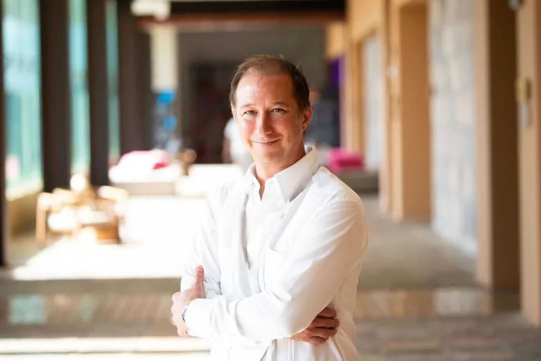 Man in white shirt stands in an indoor corridor with arms crossed, smiling at camera