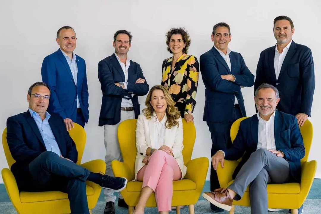 Nine business professionals pose for a group portrait, with three seated on yellow chairs and six standing behind them against a white wall