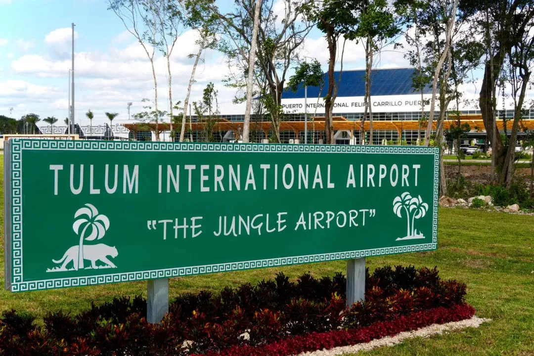 A green airport sign with palm tree decorations stands in landscaped grounds, with airport terminal buildings visible in the background