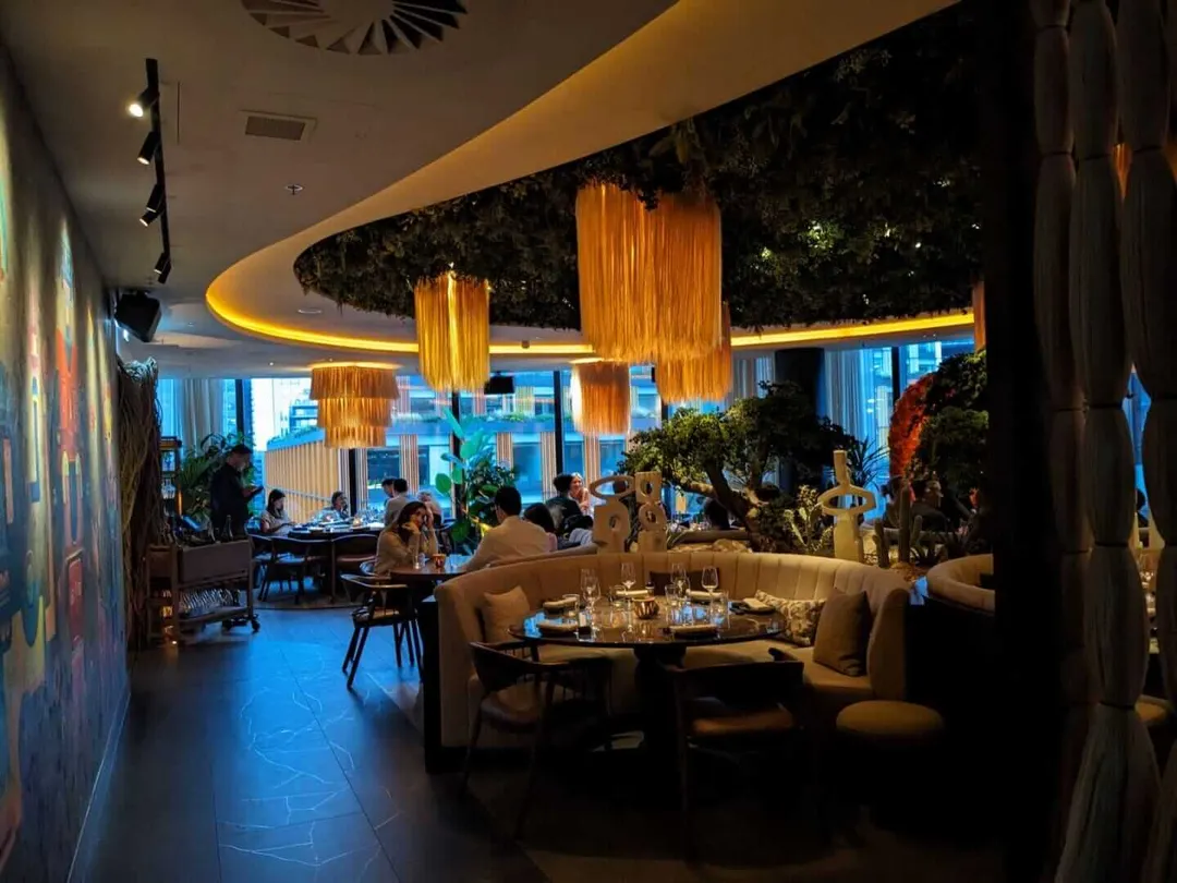 Modern restaurant interior with warm lighting, hanging golden pendant lamps, curved seating booths, potted plants, and large windows overlooking a city street a