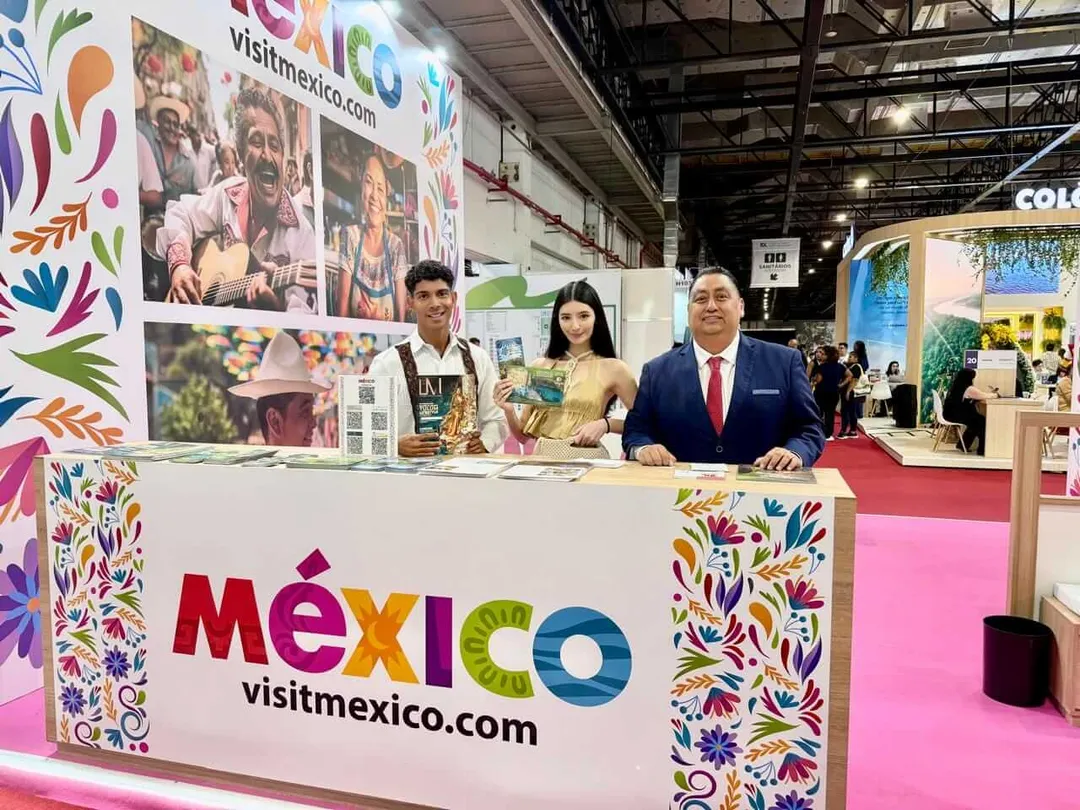 Tulum boosts winter tourism campaign in Europe with key travel expos Three people stand behind a colorful Mexico tourism booth at a travel exhibition, with promotional photos and decorative motifs displayed on the backdrop