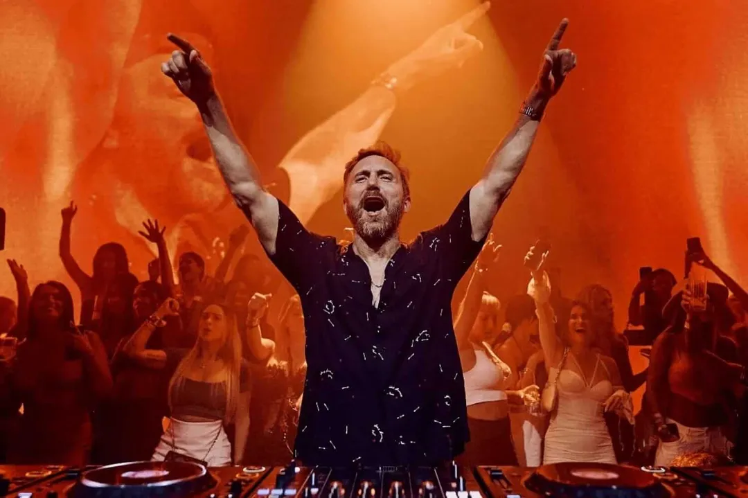 DJ at a turntable with arms raised celebrating before a crowd bathed in orange stage lighting