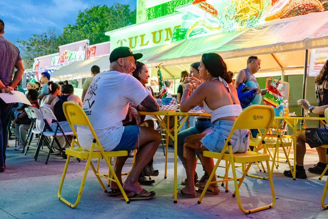 Visitors sit at yellow tables eating at an outdoor food festival with green vendor stalls in the background