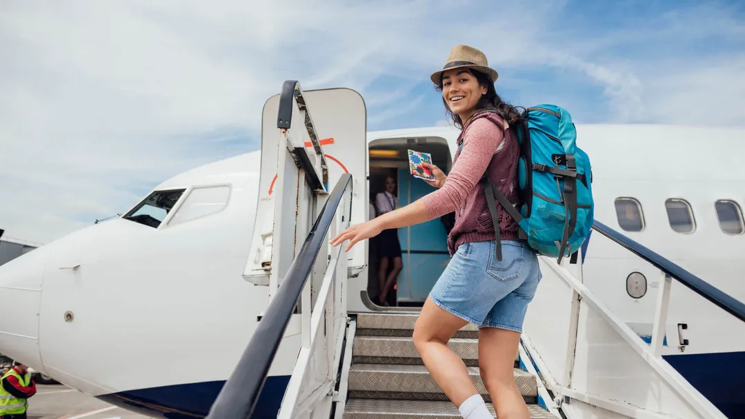 Woman wearing a hat and backpack boards an airplane via stairs, smiling at the camera