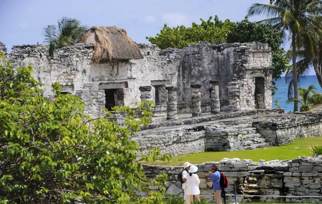 21,000 fewer tourists visit Tulum ruins in 2025 as locals protest fees Visitors walk near a weathered stone structure with a thatched roof and multiple doorways, surrounded by tropical vegetation and ocean visible in the background