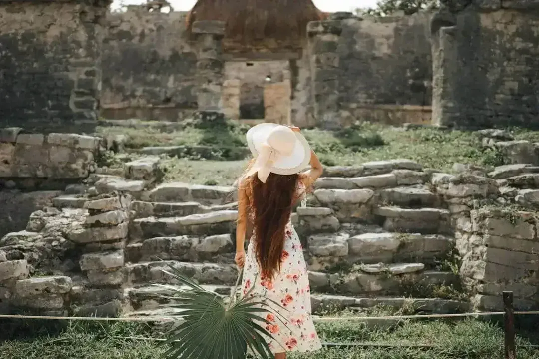 Woman in a white hat and floral dress holding a palm frond stands before ancient stone ruins