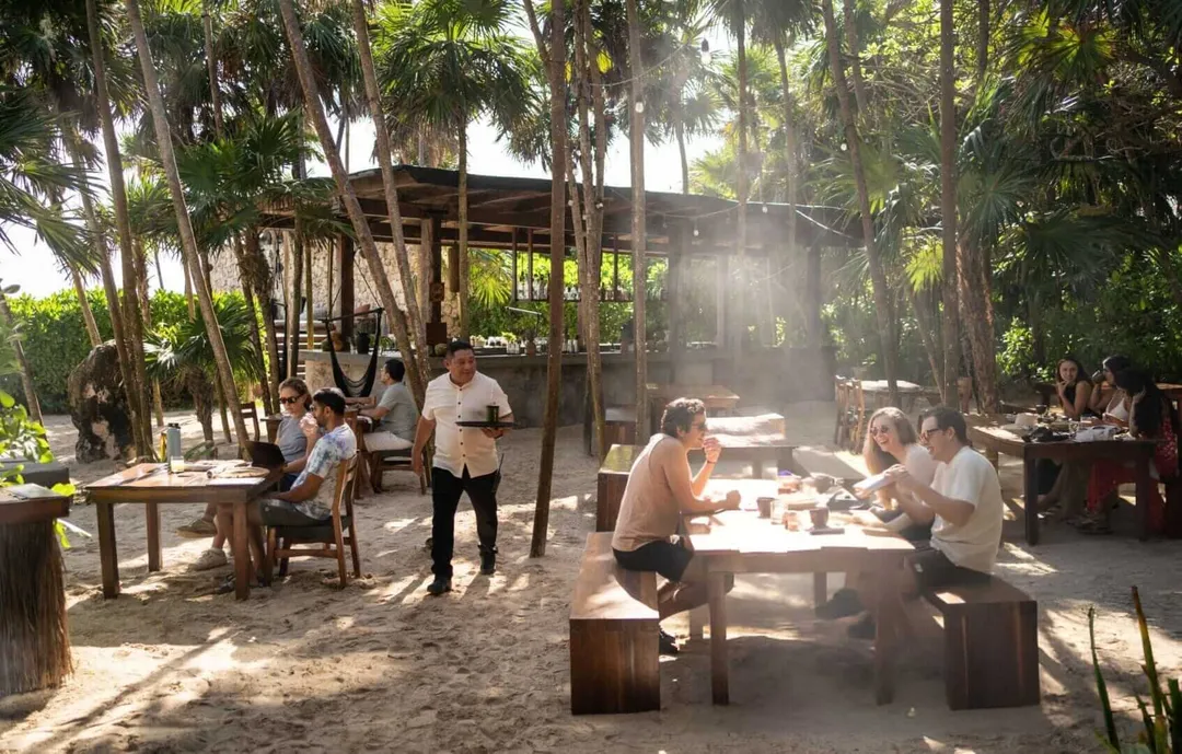 People dine and socialize at wooden tables in an open-air restaurant sheltered by palm trees and rustic structures