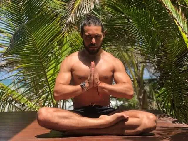 The spiritual laws Gerry Sánchez says Tulum ignored Shirtless man sits cross-legged in meditation pose beneath palm fronds with hands pressed together at chest