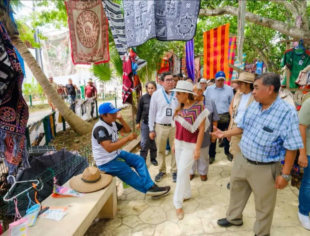 A craftsperson demonstrates traditional weaving at an outdoor market beneath palm trees while visitors browse handmade textiles and goods nearby