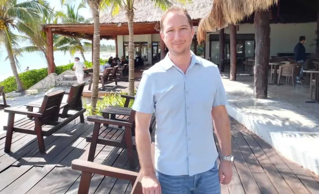 Tulum’s tourism dream runs into physical limits Man in light blue shirt standing on a wooden deck at a beachfront resort with palm trees, thatched structures and other guests visible in the background
