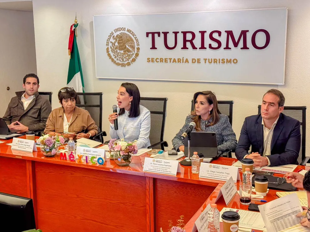 Five people sit at a red table during a panel discussion in front of a tourism ministry sign with a Mexican flag visible in the background