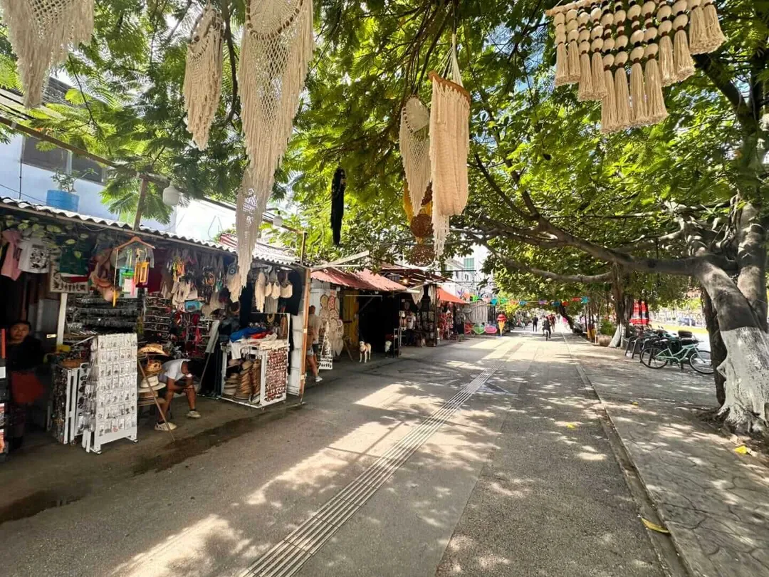 Outdoor market street lined with vendor stalls beneath leafy trees, with decorative woven ornaments hanging overhead