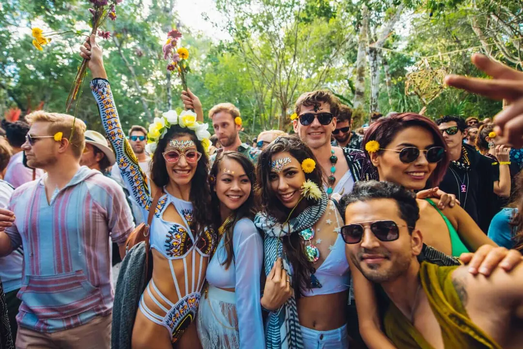 Group of people wearing sunglasses and yellow flower crowns celebrating in a wooded area with decorative body paint and accessories