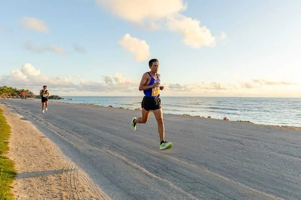 Runner wearing a race bib jogs along a sandy beach with other participants and ocean in the background