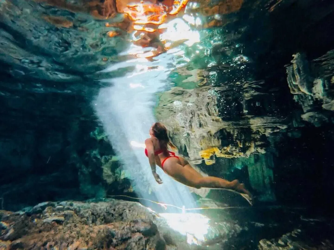 Snorkeler swimming through an underwater cenote cave with sunlight filtering down from above