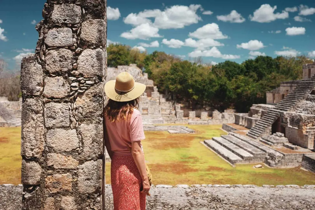 Tourism in Mexico faces fiscal pressure as investors call for reform Person in a hat and pink shirt standing beside a stone column at an ancient ruins site with pyramids and forest visible in the background