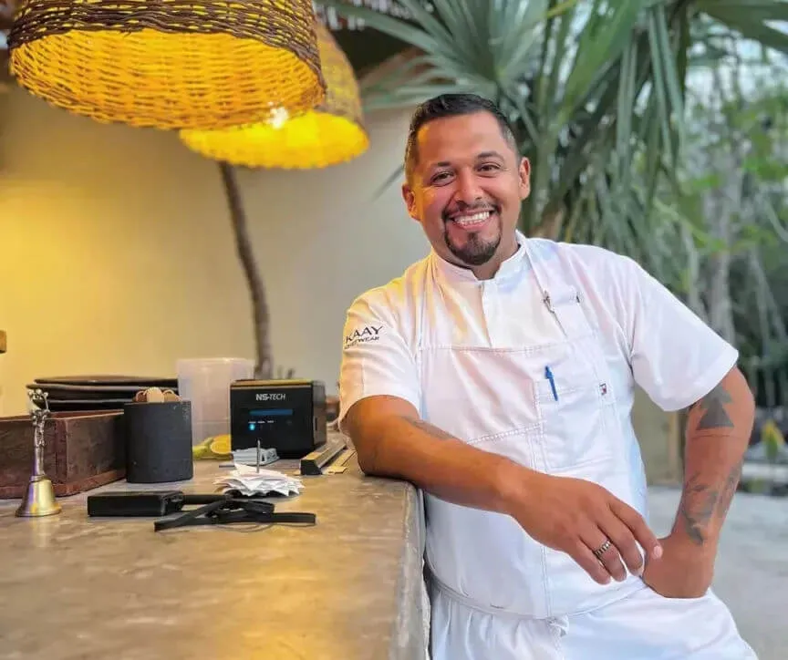 Chef in white uniform smiles while leaning against a counter in a modern restaurant space with plants and woven pendant lighting