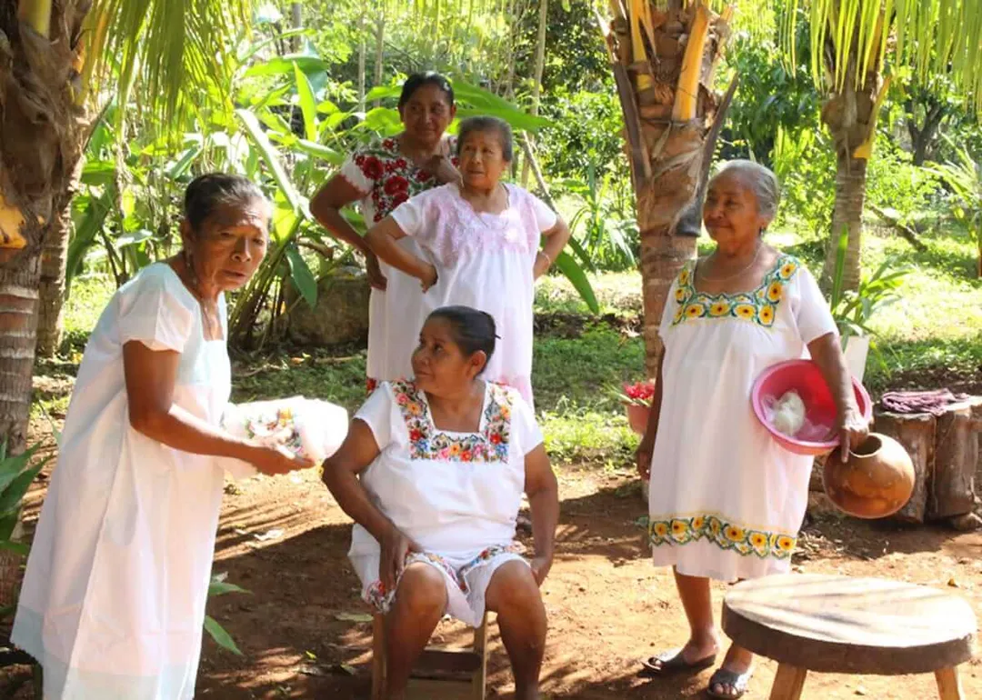 Five women in white embroidered dresses gather in a tropical garden setting with palm trees and basic wooden structures visible in the background