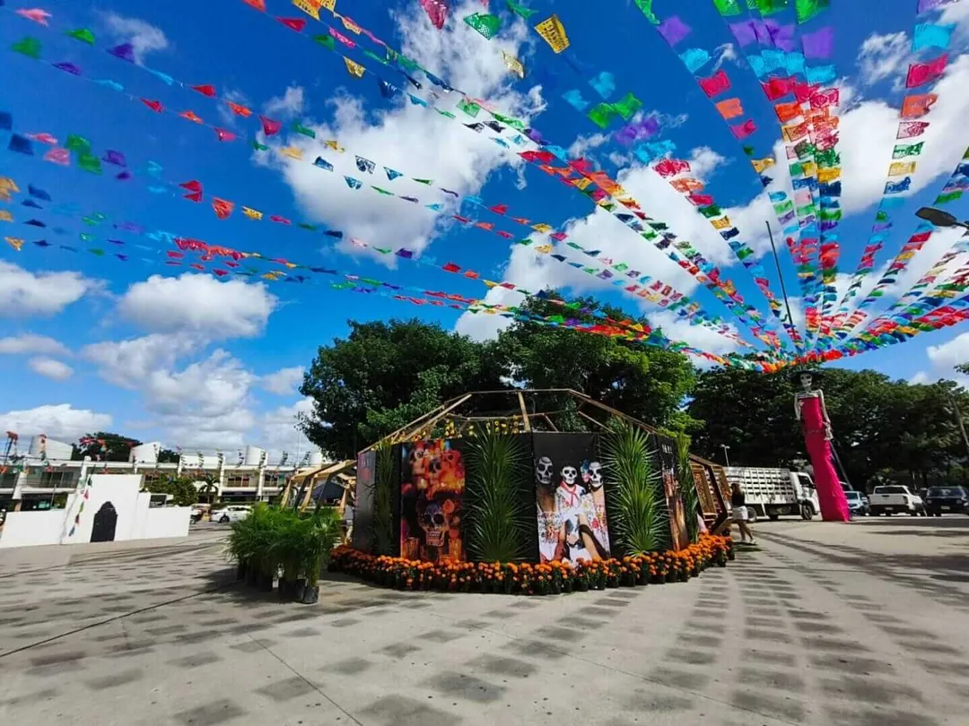 Colorful papel picado banners hang above a decorated wooden structure adorned with skull imagery and marigolds in an outdoor plaza