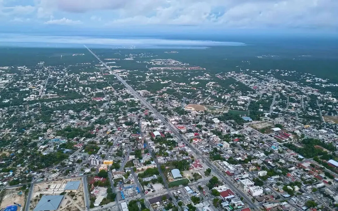 Real estate leaders push for larger, affordable homes in Tulum Aerial view of a coastal town with a straight highway running through residential areas, ocean visible in the distance under partly cloudy sky