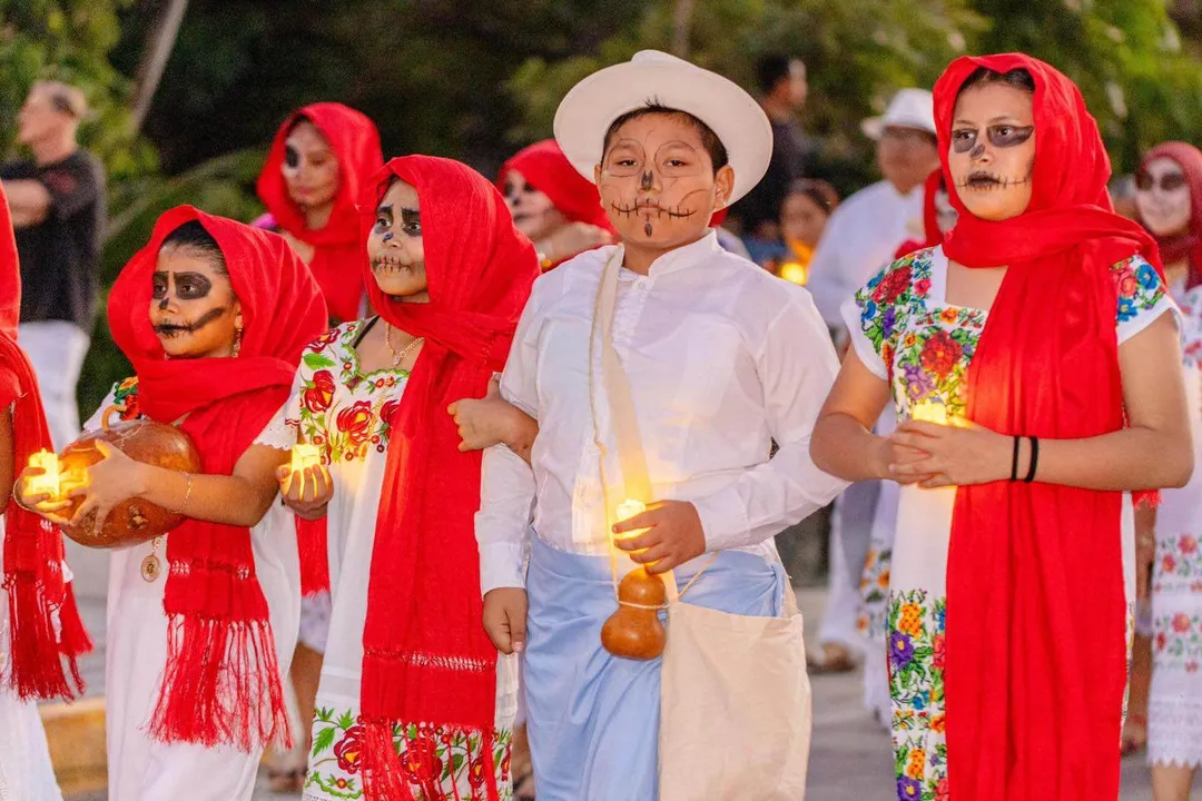 Children wearing skeleton makeup and red clothing hold lit candles during an outdoor nighttime procession