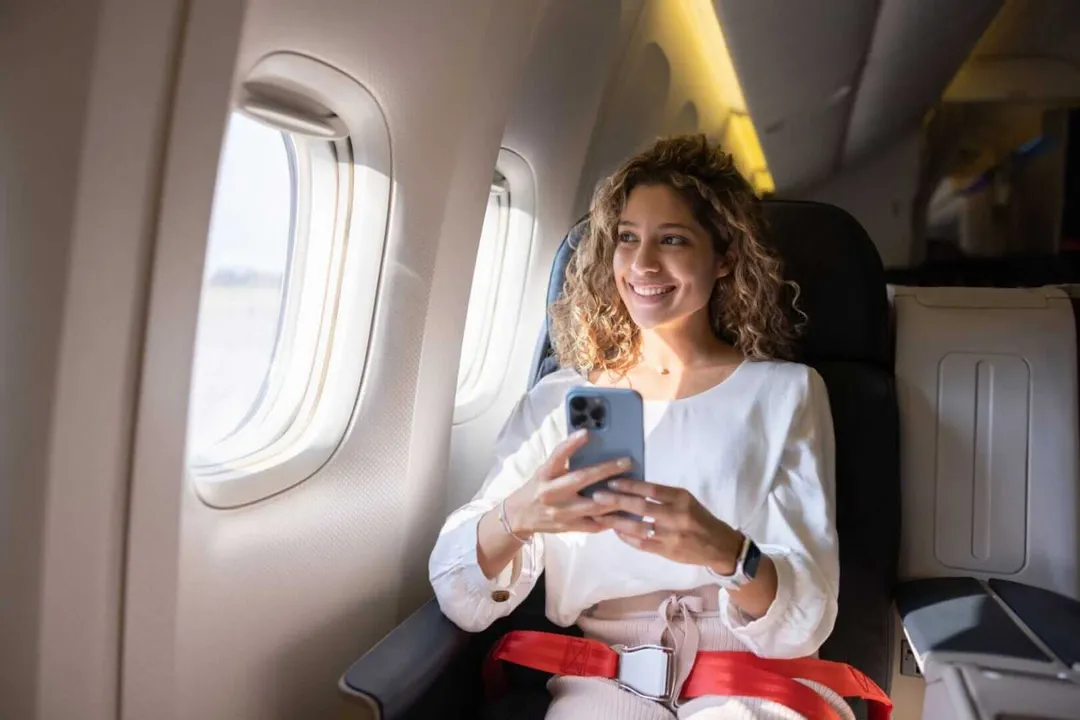 Woman seated in an airplane window seat holding a smartphone and smiling