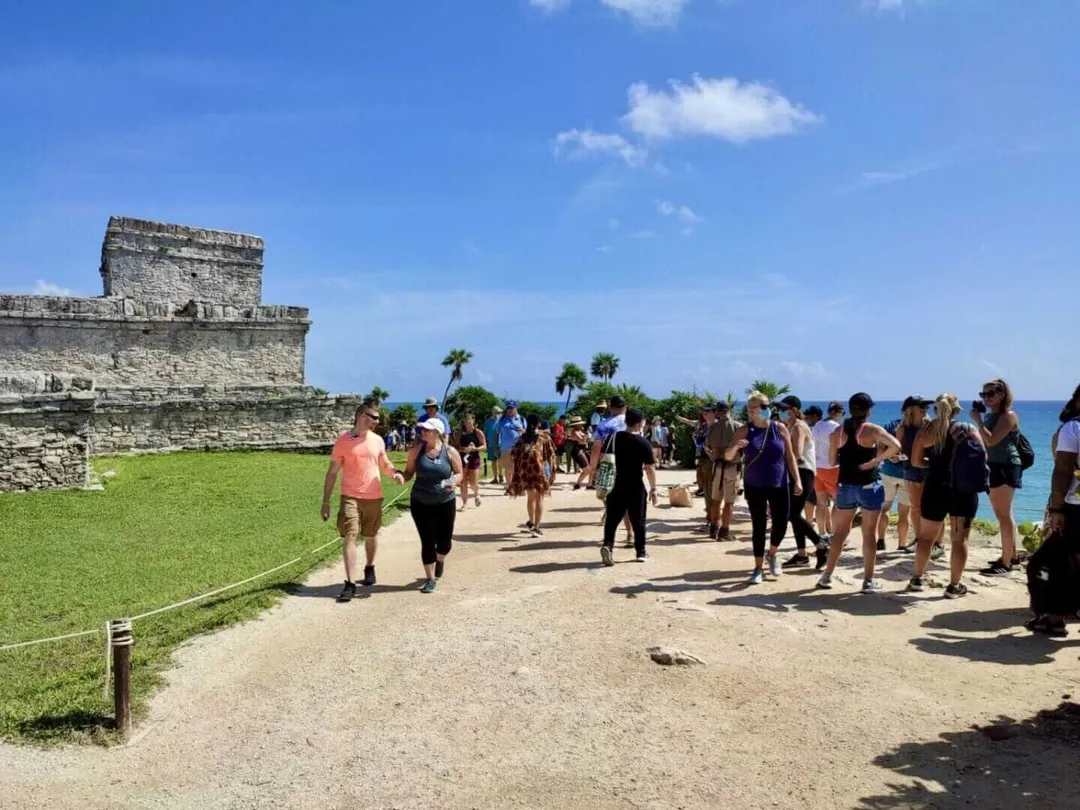 Tourists gather at a beachfront archaeological site with a stone structure, palm trees, and ocean visible in the background
