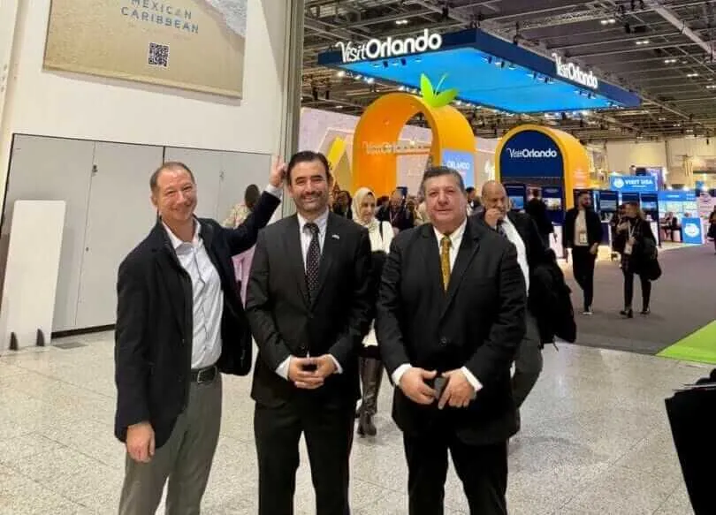 Three men in business suits stand together at a convention center with a large orange tourism logo and "Visit Orlando" signage visible behind them