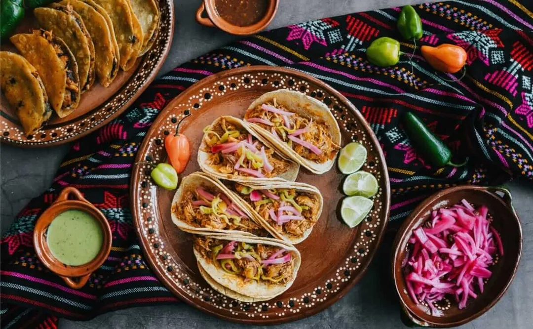 Four tacos filled with shredded meat and cabbage sit on a decorative plate surrounded by lime slices, with fried tortillas, sauces, and pickled vegetables arran