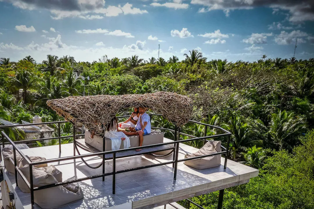 Tulum vacation rentals show rising supply and falling demand Two people sit at a table on an elevated wooden deck overlooking dense tropical vegetation and palm trees under a partly cloudy sky