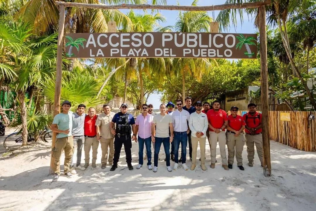 La playa del pueblo reveals what Tulum never said A group of men stand in a line beneath a wooden archway sign on a sandy beach lined with palm trees