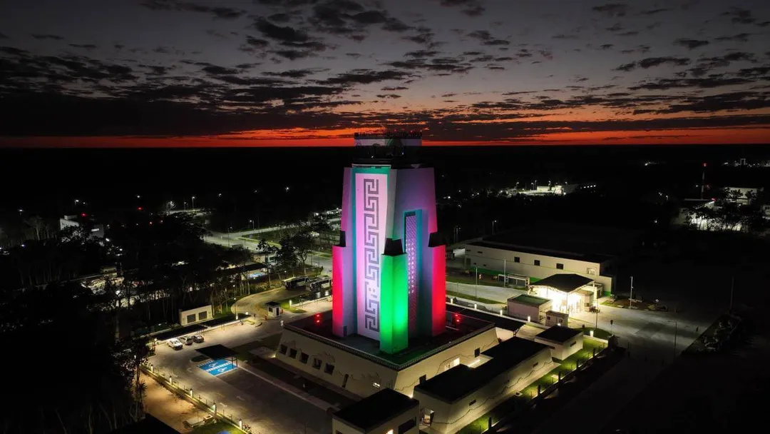 An illuminated geometric structure with pink, purple and green lighting sits atop a building complex at dusk, surrounded by other lit structures and darkened la