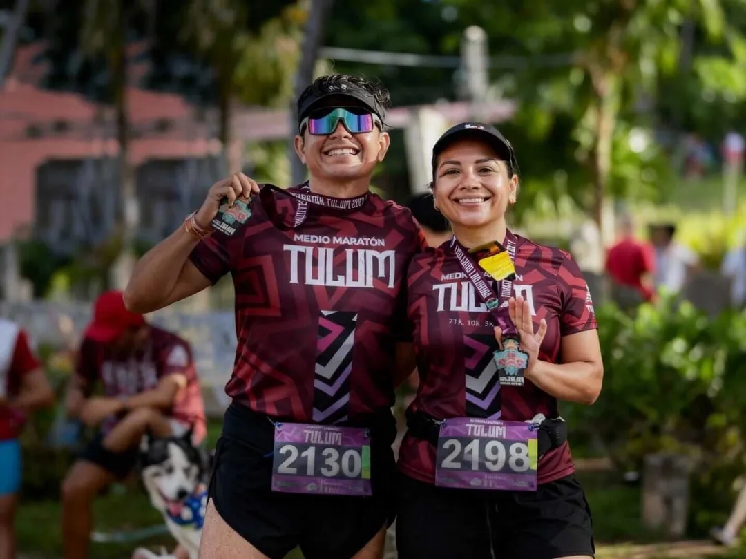 Two marathon runners in matching maroon race bibs display their medals while smiling at the finish line, with spectators and green trees visible in the backgrou