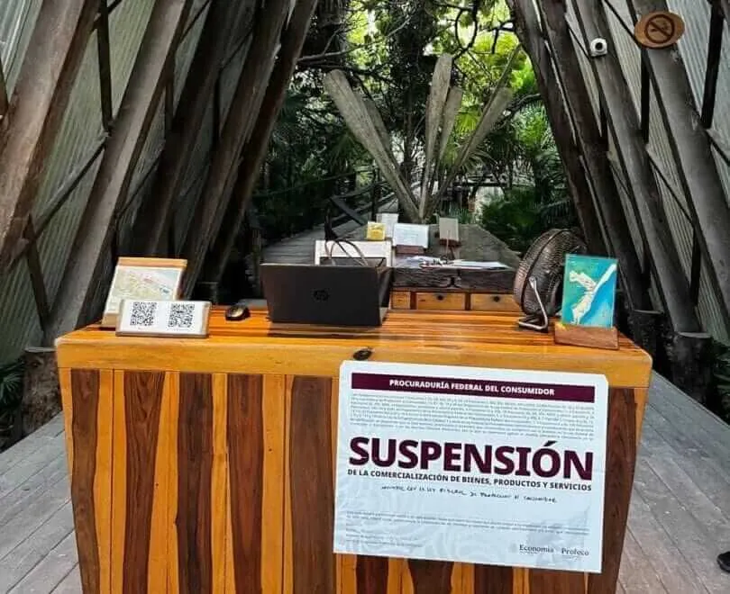 A wooden desk with a laptop and informational materials sits under a tent structure in a tree-lined outdoor setting, with a suspension notice posted on the fron