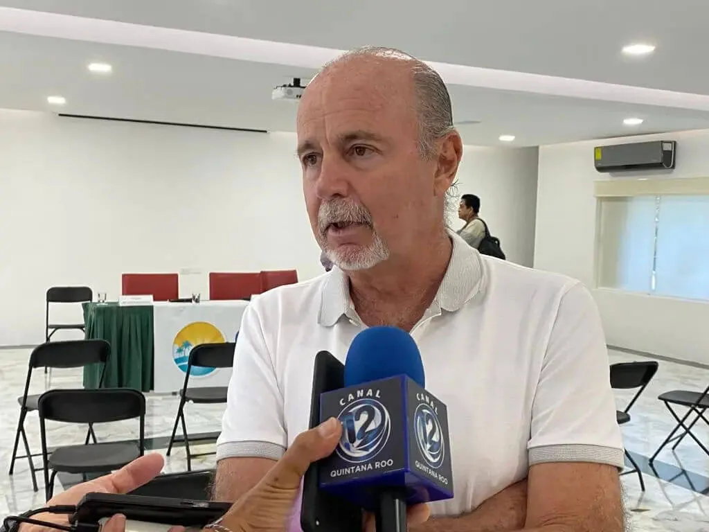 Hotel executive denounces local online attacks on Tulum Man in white polo shirt speaking into microphones in an indoor meeting room with folding chairs and a banner visible in the background