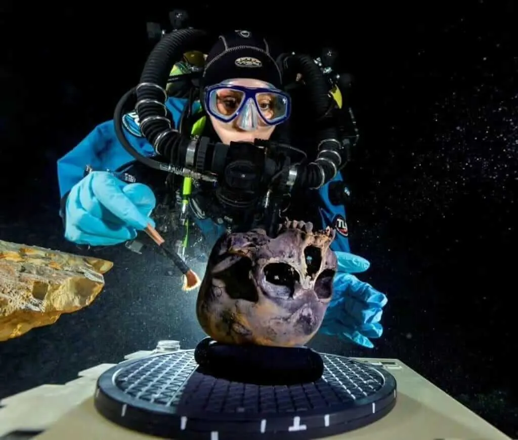 Scuba diver in blue wetsuit and mask examines a skull artifact on the seafloor underwater