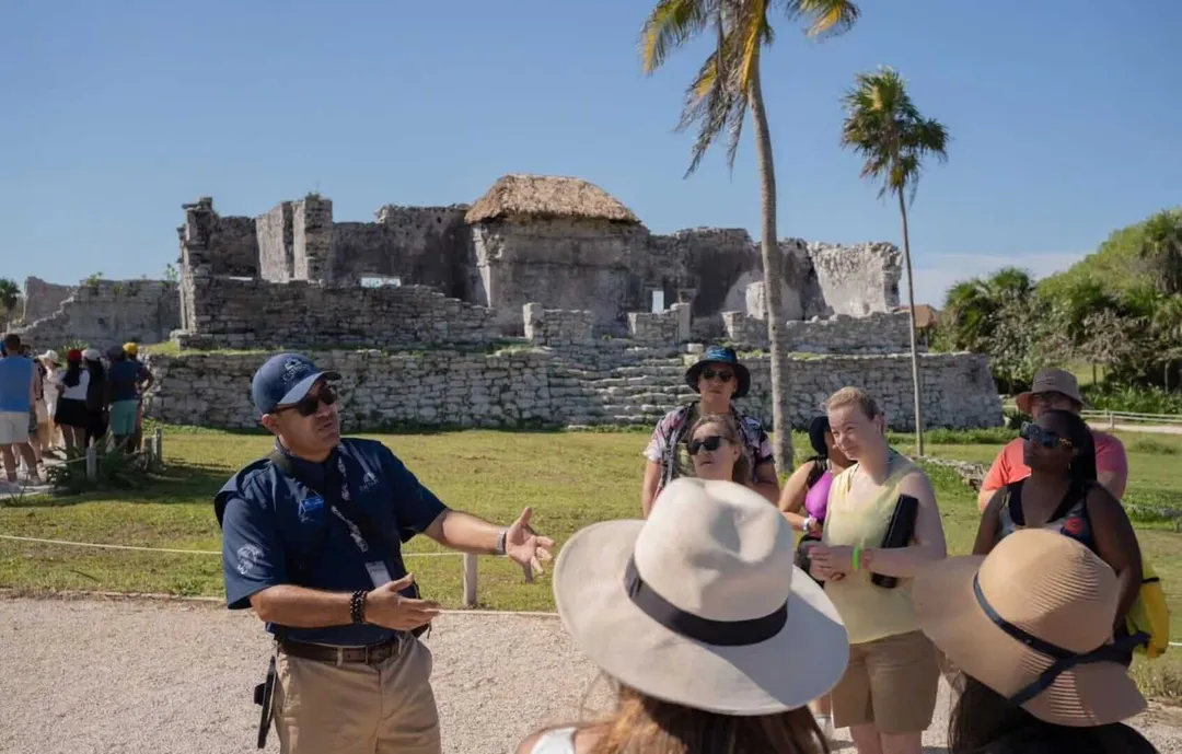 Economy in Tulum grows cautiously amid weaker US arrivals A tour guide speaks to a group of visitors in front of ancient stone ruins with palm trees in a grassy setting