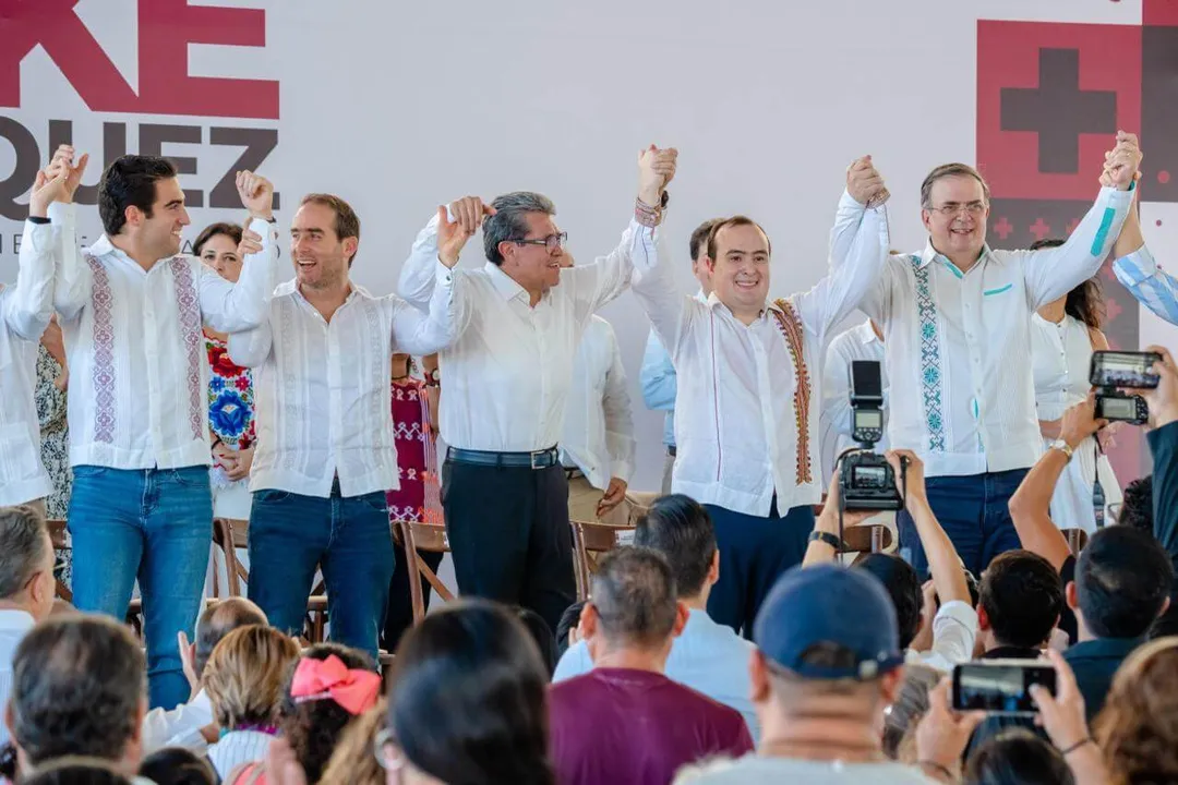 Several men in white guayaberas and blue pants stand on stage with raised joined hands while a crowd below photographs them