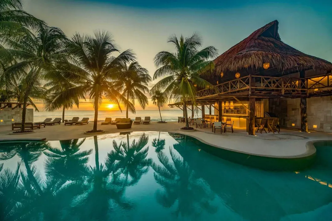 Resort pool with thatched-roof palapa, palm trees, and beach cabanas at sunset overlooking the ocean