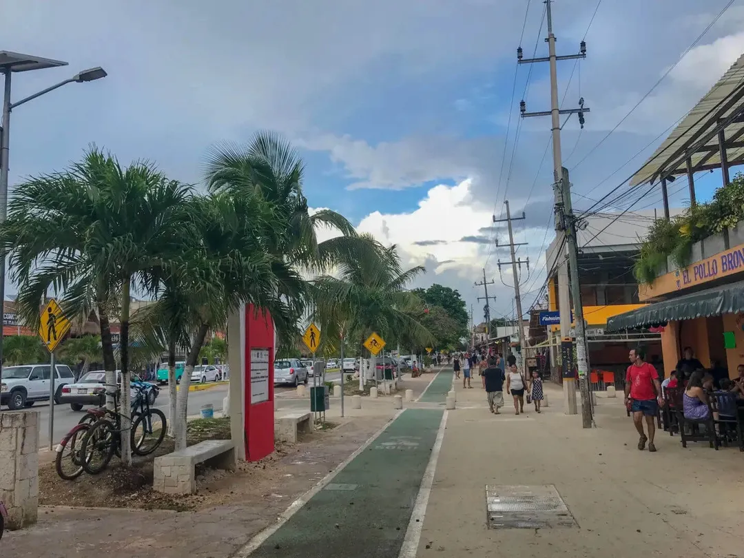 Pedestrians walk along a tree-lined street with commercial storefronts, parked vehicles, and power lines in a tropical town setting