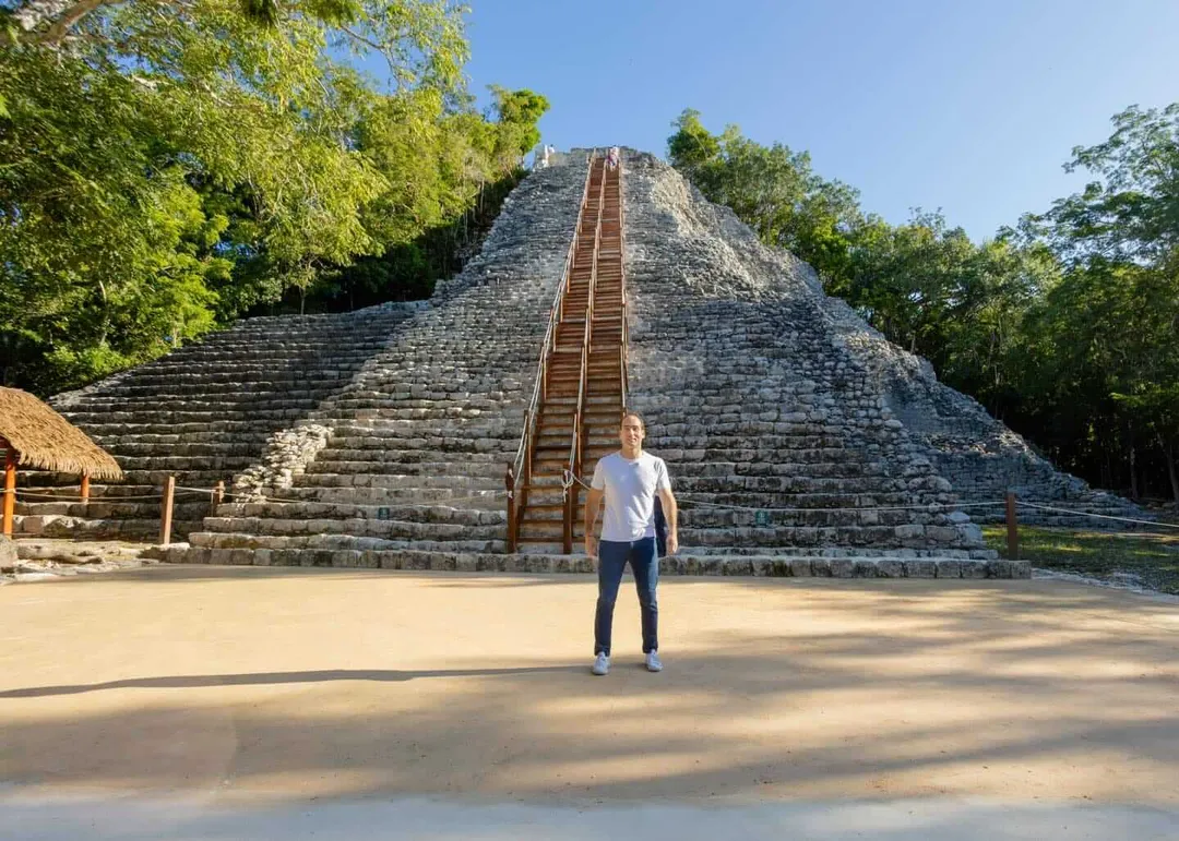 A person stands in front of a large stone pyramid with a wooden staircase running up its center, surrounded by jungle vegetation under clear blue sky
