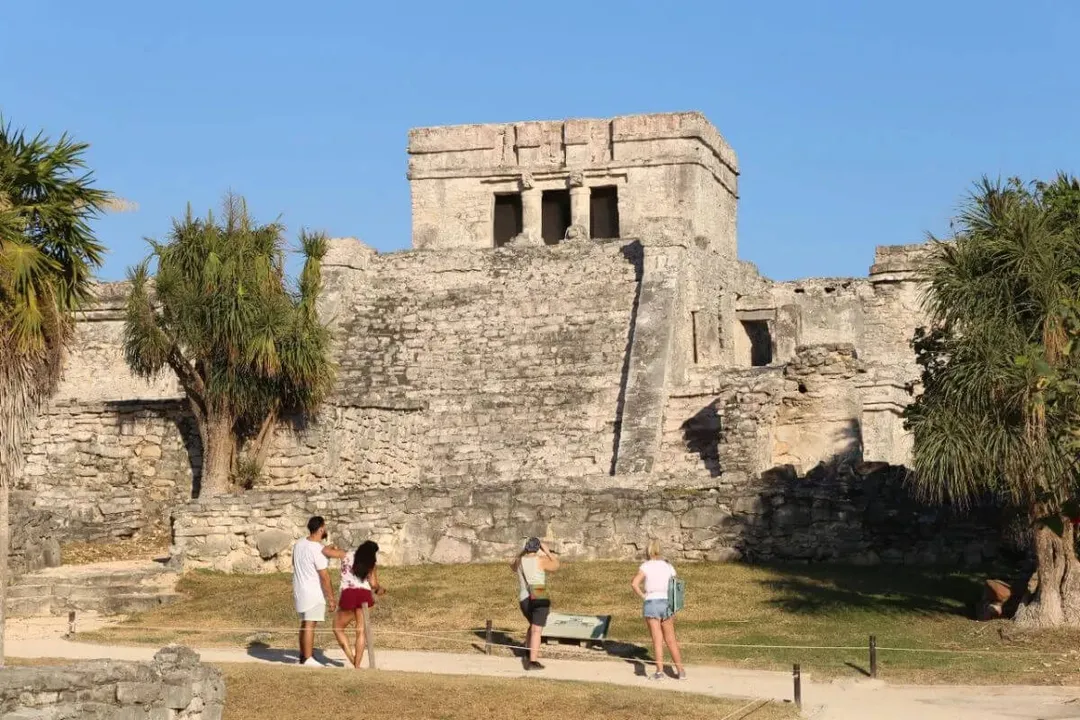 Tulum Archaeological Zone Remains Mexico’s Third Most Visited Site Visitors stand in a grassy courtyard facing a large stone structure with rectangular windows and palm trees on either side under clear blue sky