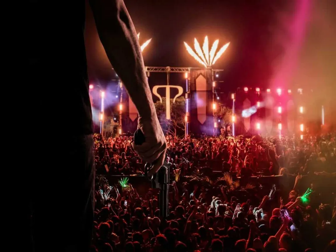 A large crowd gathers at night in front of an illuminated stage structure with colorful lighting and decorative elements overhead