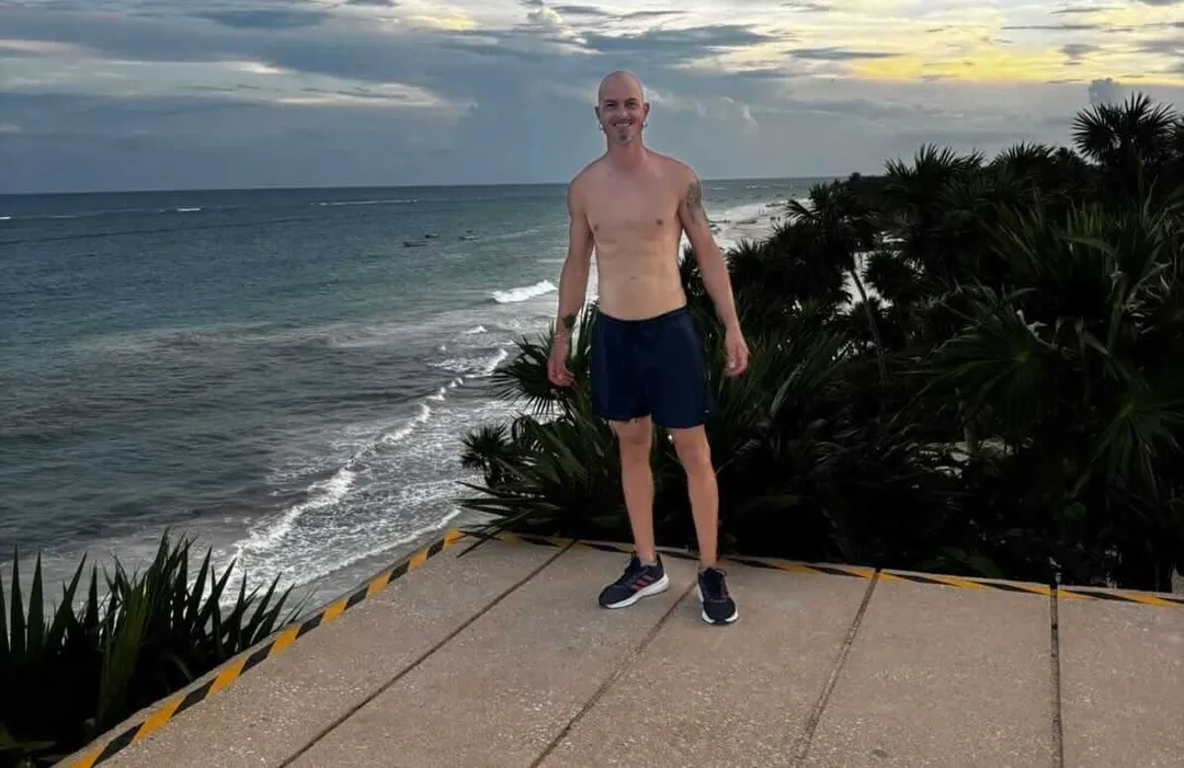 Shirtless man standing on a concrete platform overlooking a beach with palm trees and ocean waves at dusk