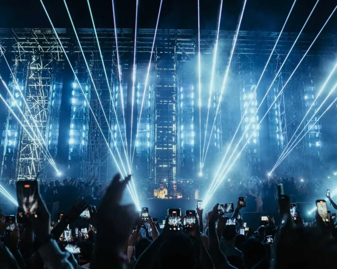 Crowd at an indoor electronic music concert with dramatic blue stage lighting beaming upward from the performance area