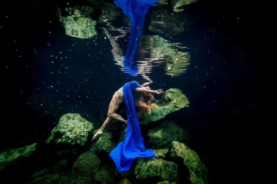 A person in a blue fabric performs an underwater movement among moss-covered rocks in a dark cenote or cave pool
