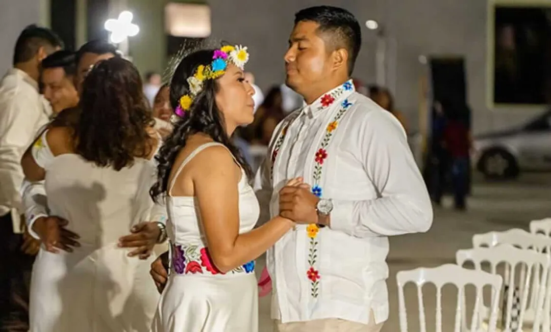 Tulum opens registration for free collective weddings in 2026 A couple wearing white clothing and colorful flower garlands dance together at an outdoor evening celebration with other guests in the background