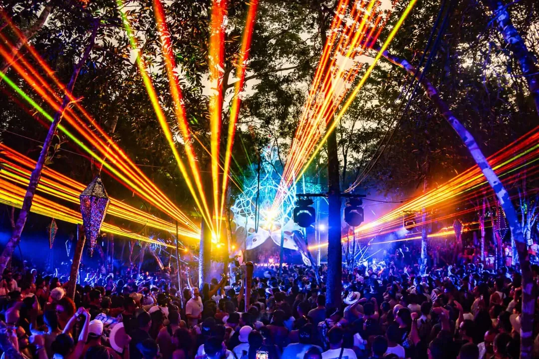 Day Zero Tulum 2026 Marks Thirteen Years of Music, Ritual, and Community Crowds of people gather under trees at night beneath elaborate red, orange, yellow and blue laser light beams radiating from a central stage area