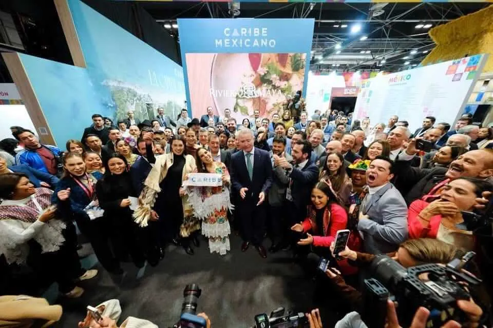 A large crowd gathers around officials and a person in traditional dress at an indoor tourism exhibition booth labeled "Caribe Mexicano