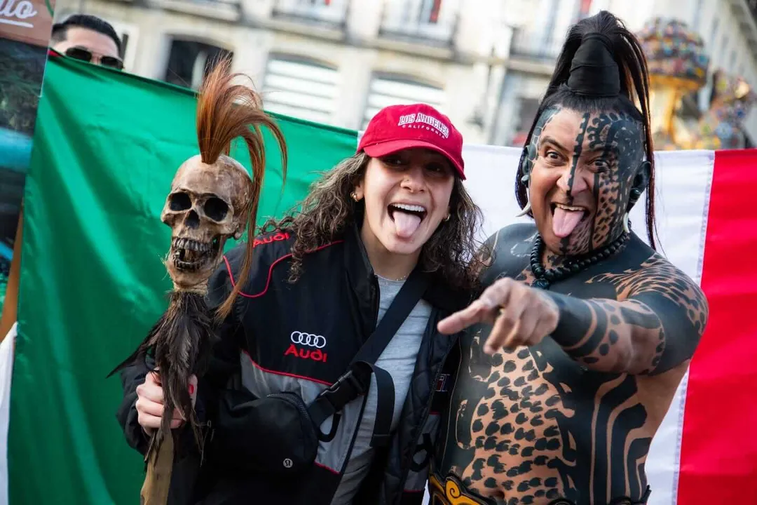 Two people in costume pose with a skeleton prop while holding a Mexican flag in an urban setting